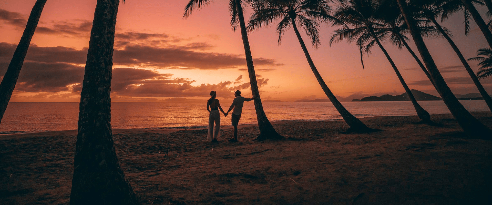 Couple Standing Amongst Palm Trees On Beach During Sunset